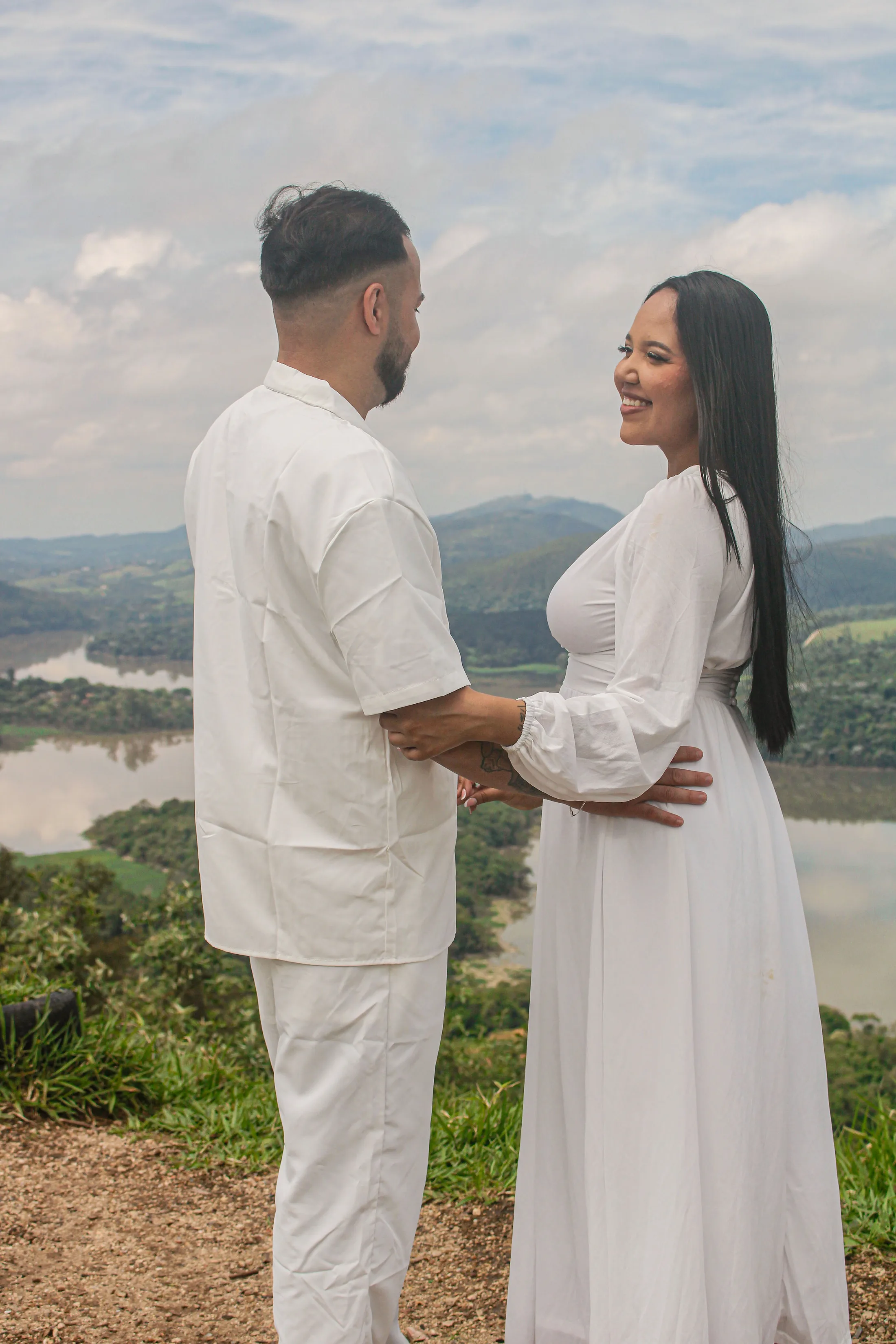 Casal de roupas brancas frente a frente, sorrindo, em um mirante com represa e montanhas ao fundo.