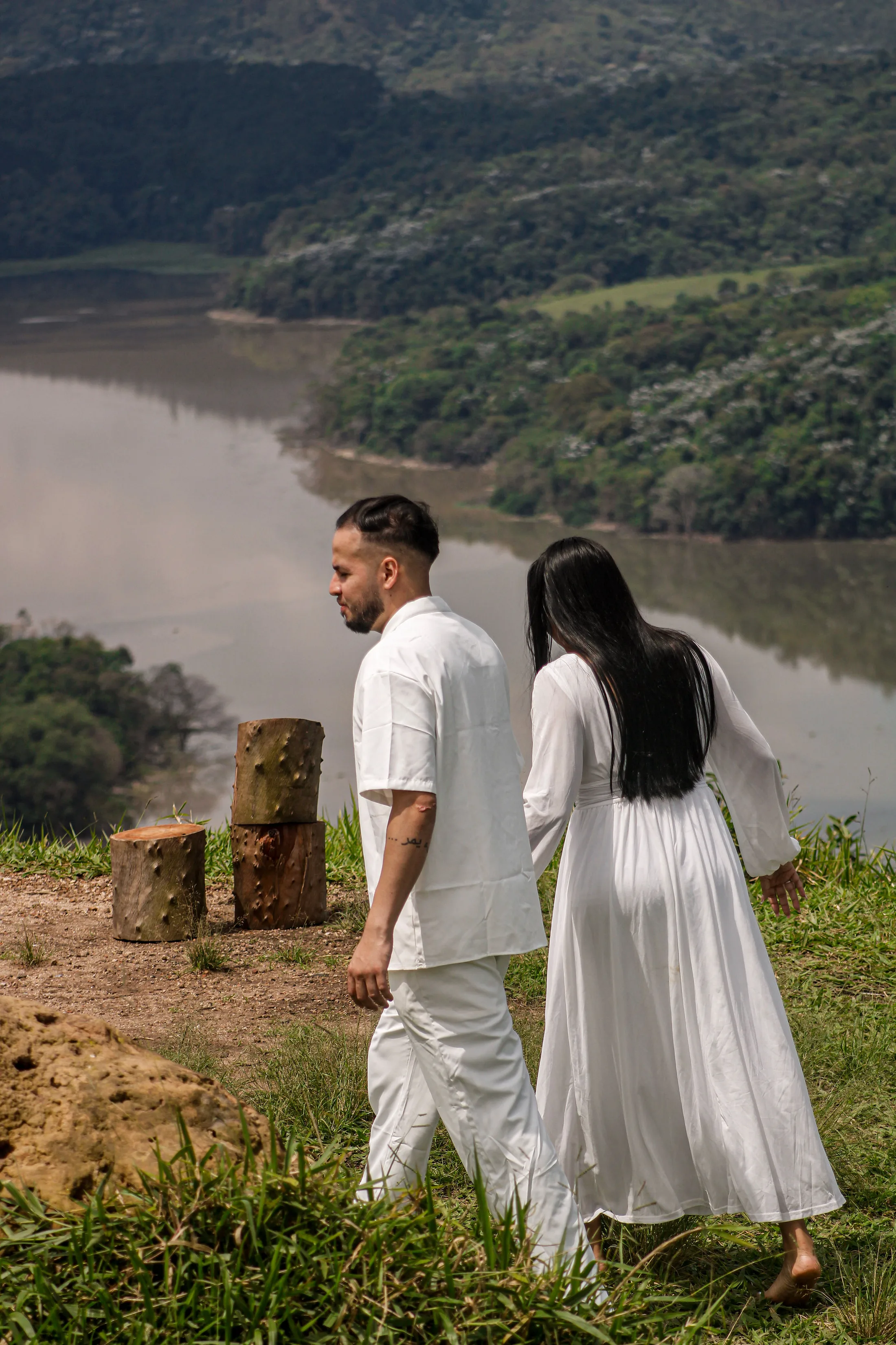 Casal de roupas brancas caminhando de costas em direção à represa, em um mirante.