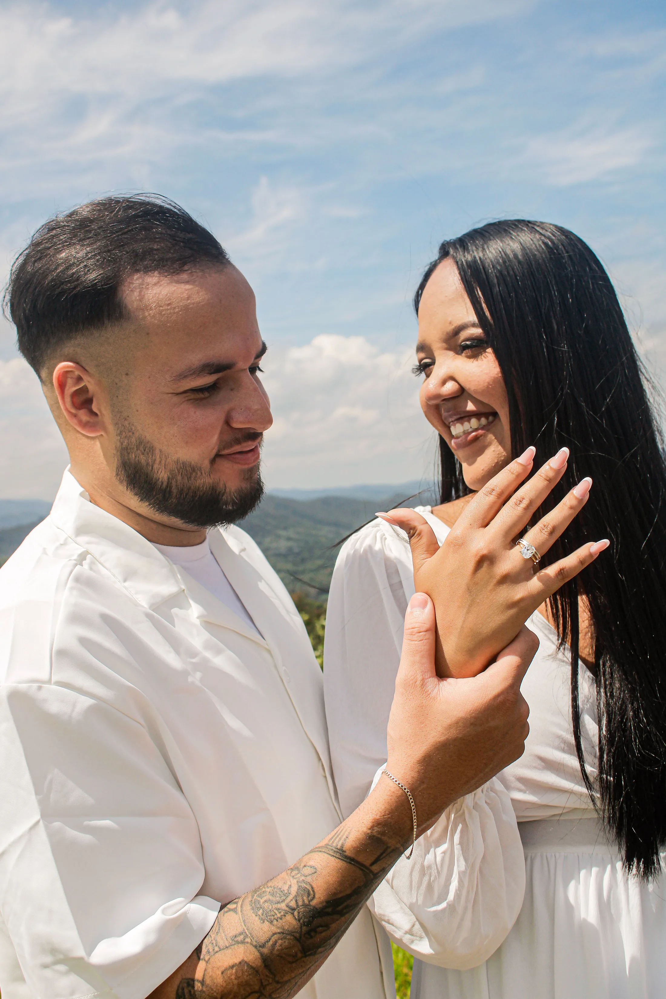 Casal sorrindo com as mãos unidas; ela usa um anel.