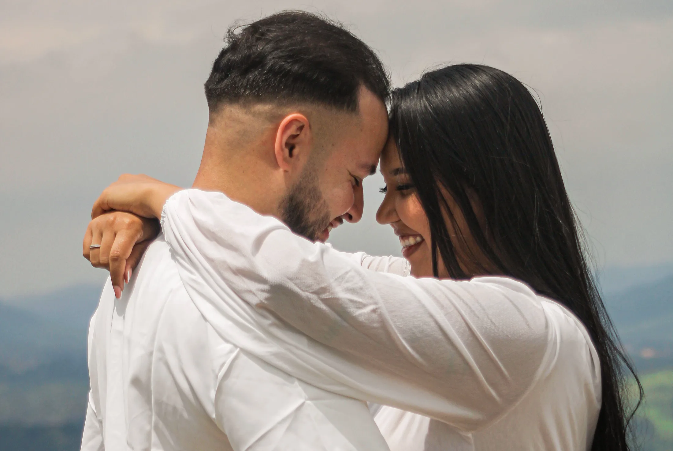 Casal com as testas encostadas, em close, com montanhas ao fundo.
