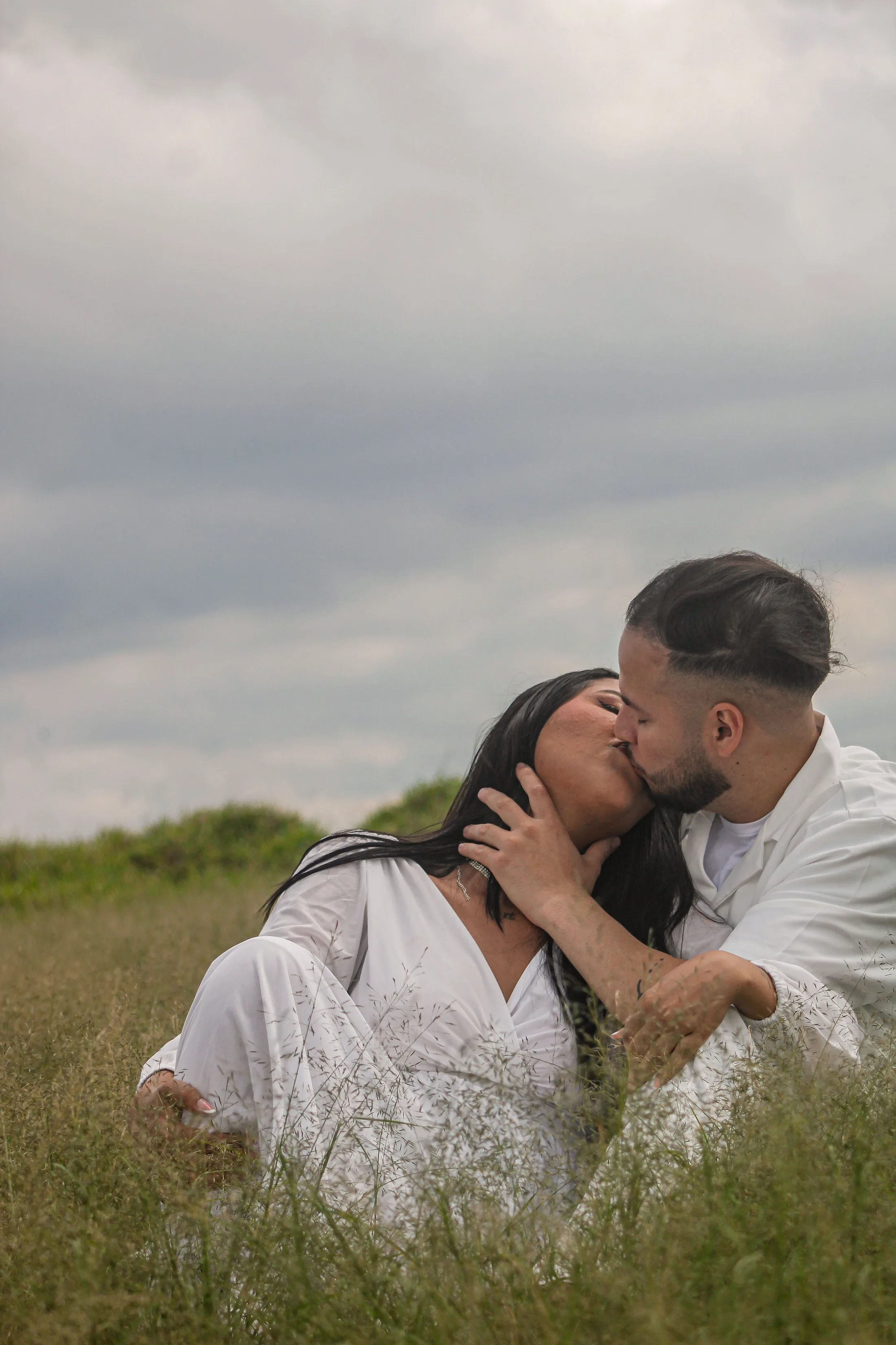 Casal se beijando na grama, com céu nublado ao fundo.