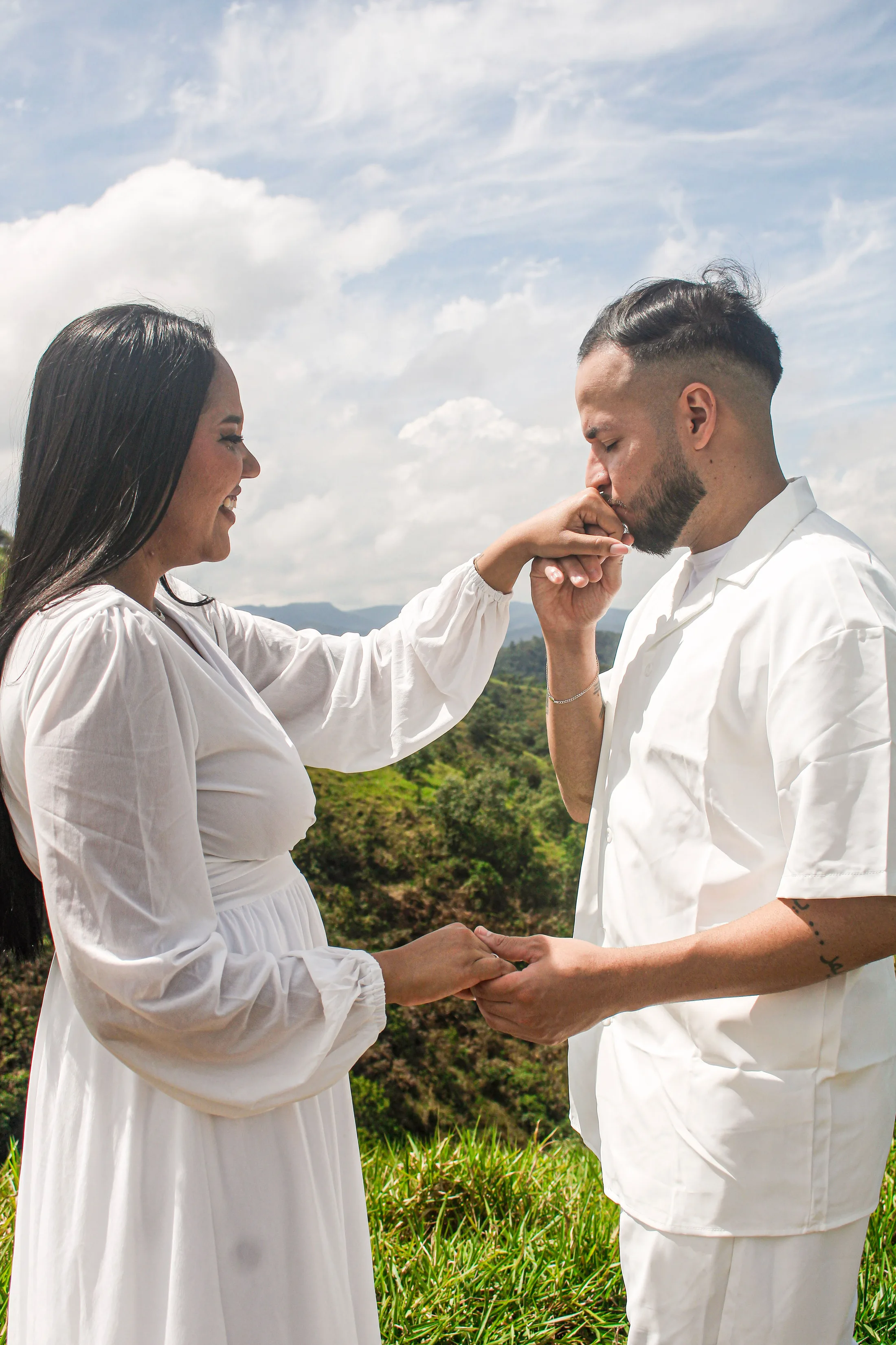 Homem beijando a mão da mulher, que usa um anel, com montanhas ao fundo.