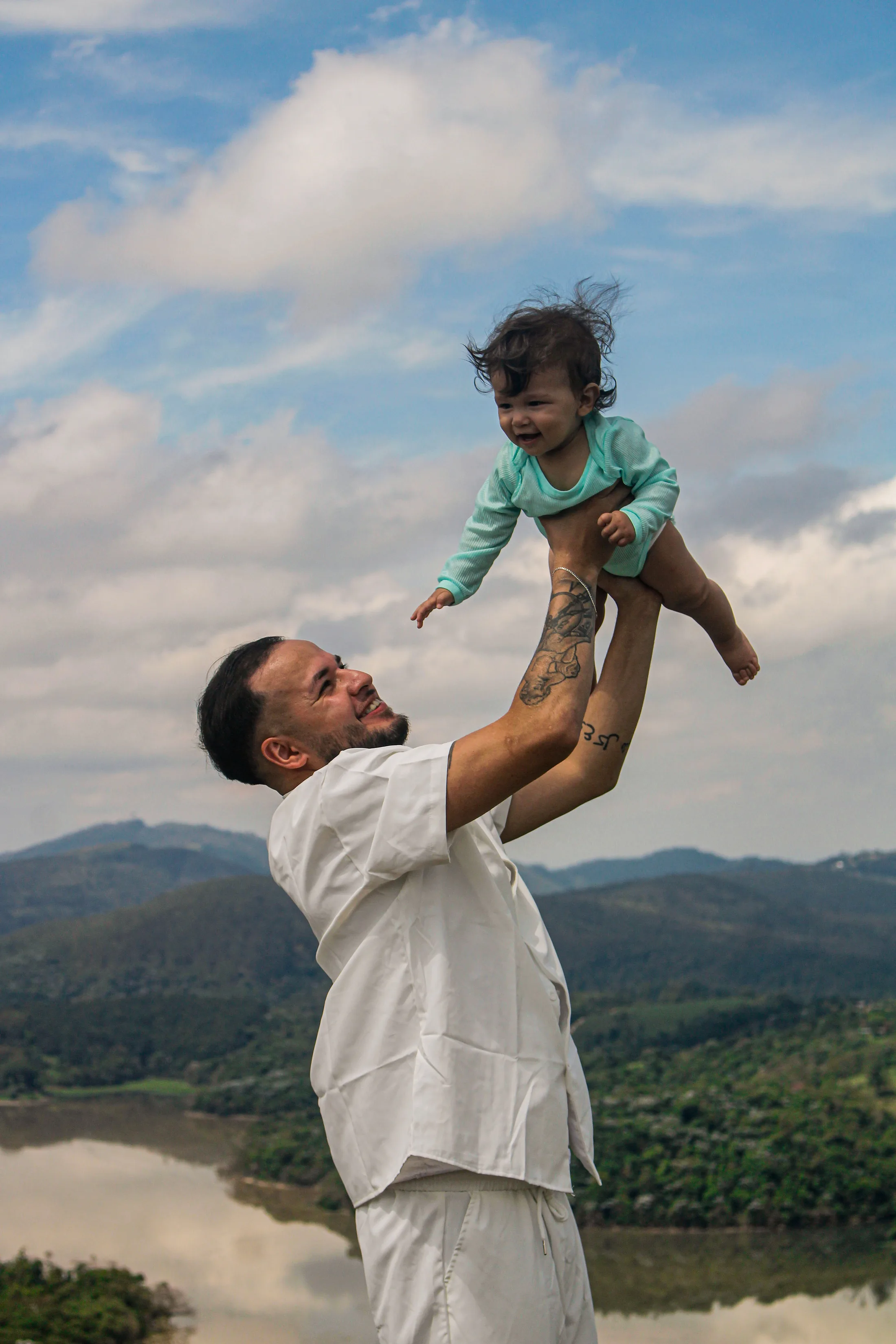 Homem erguendo uma criança no ar, com céu azul e a represa ao fundo.
