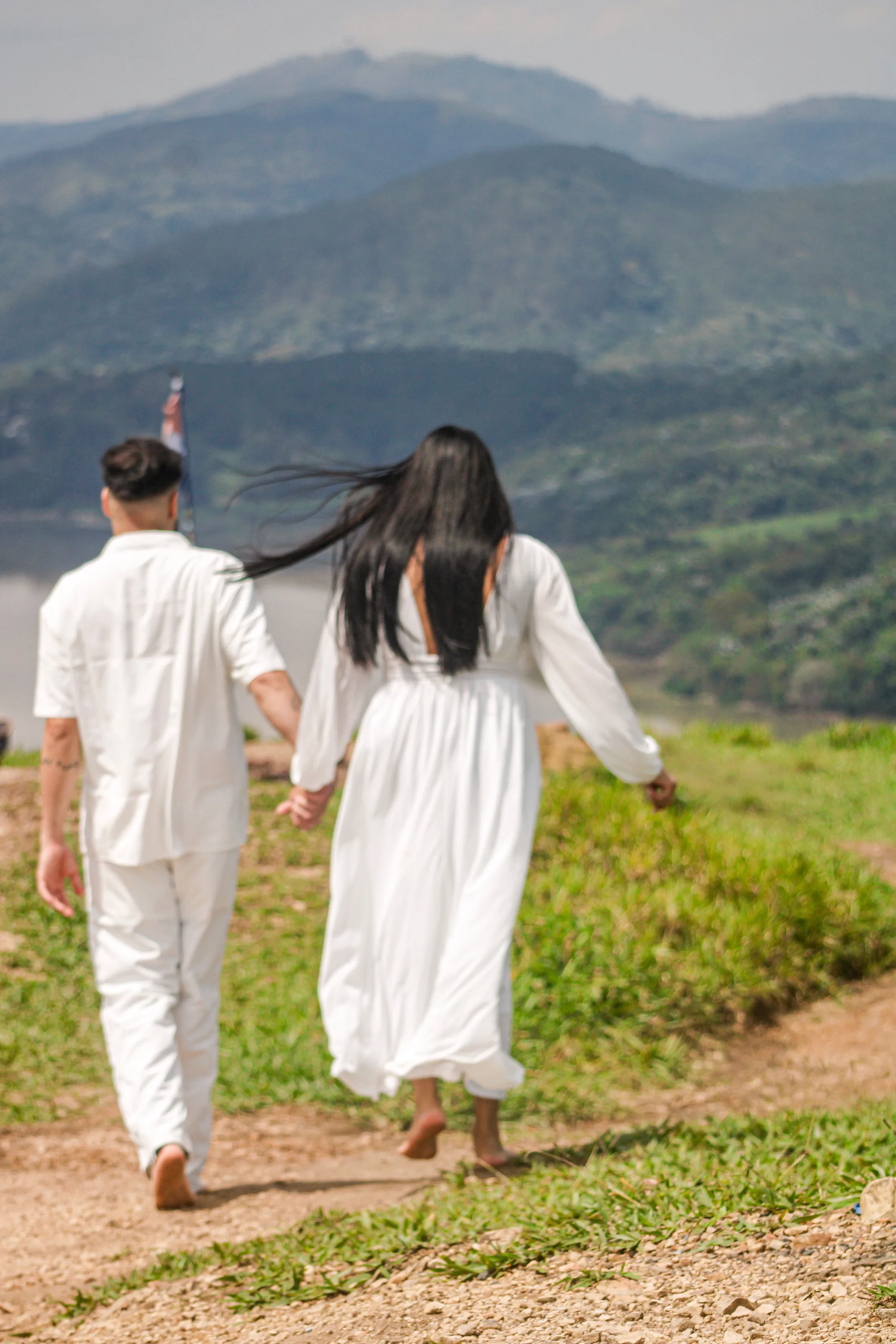 Casal caminhando de costas por uma trilha de terra, com montanhas ao fundo.