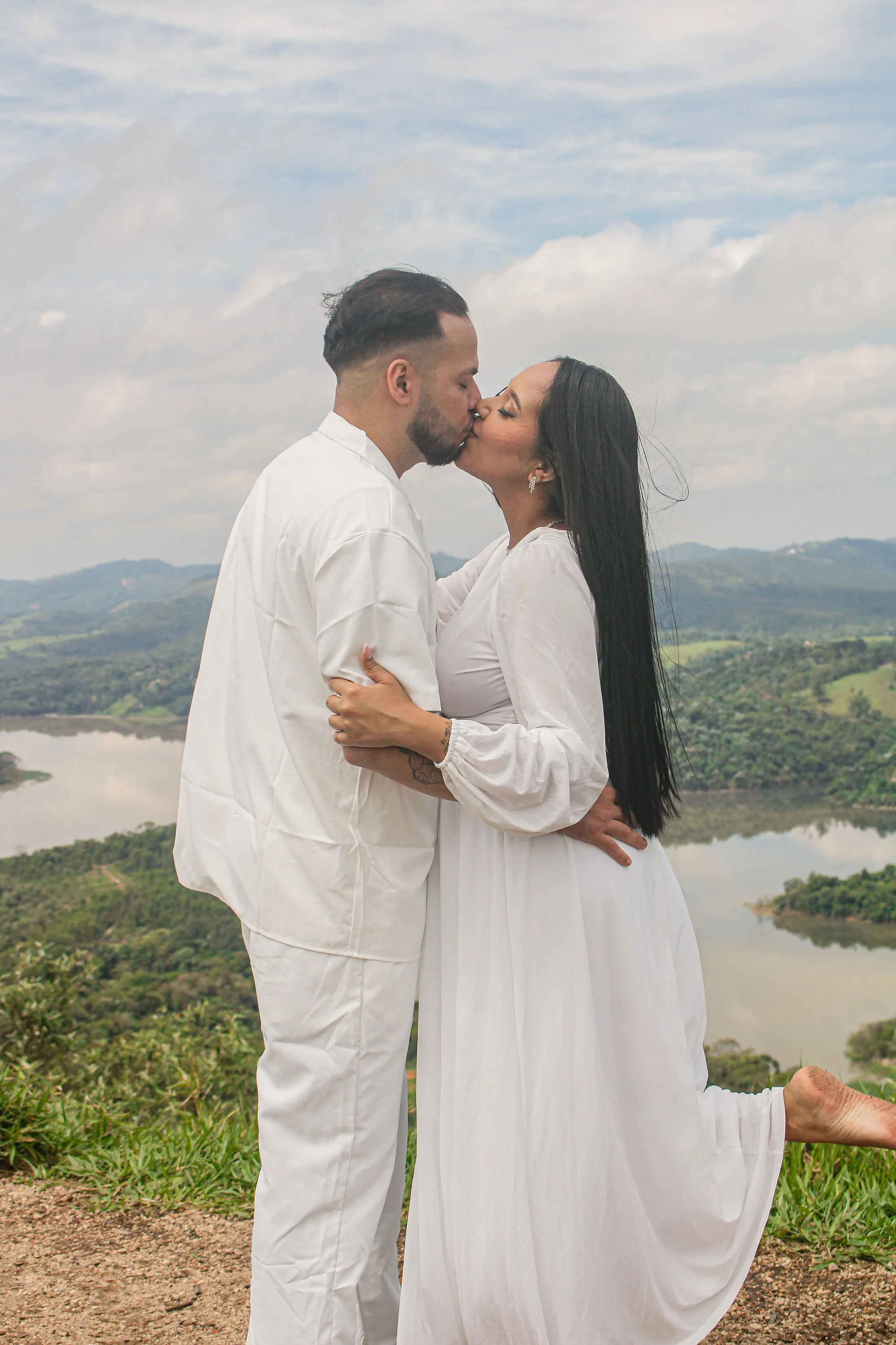 Casal de roupas brancas se beijando em um mirante, com a represa ao fundo.