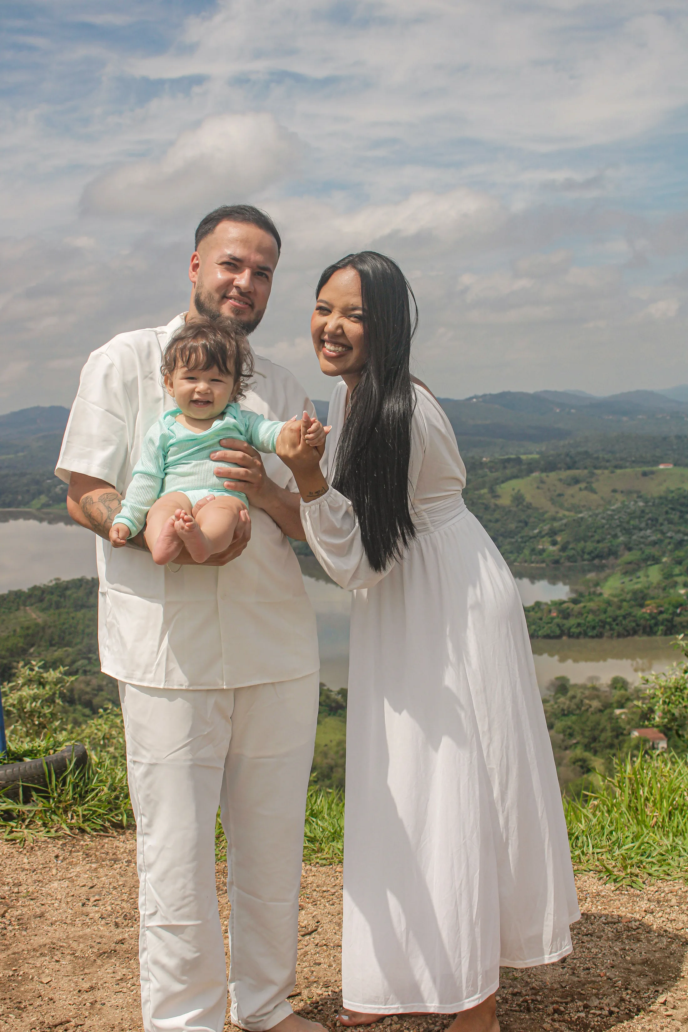 Casal com uma criança no colo, sorrindo em um mirante com a represa ao fundo.