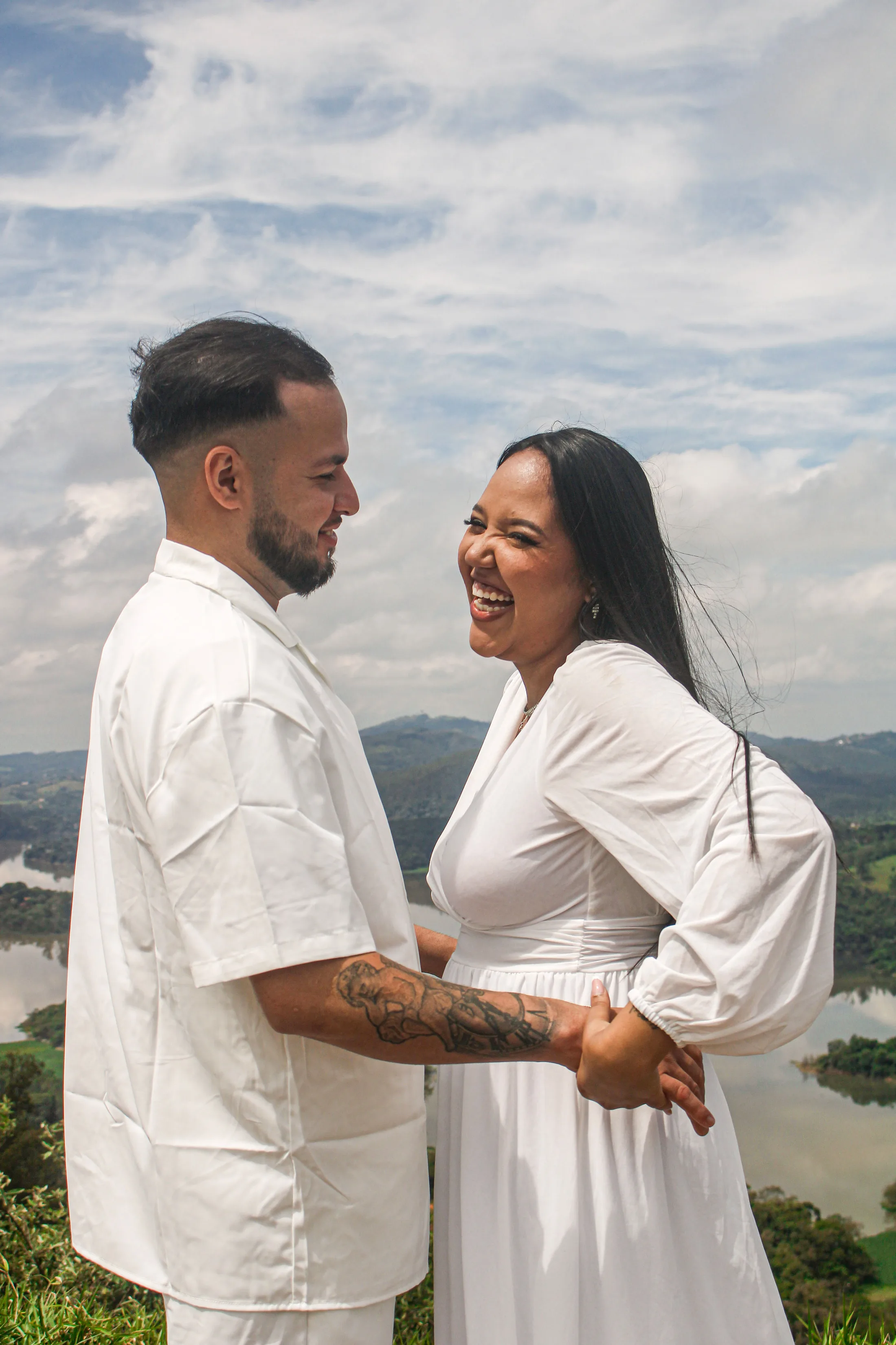 Casal rindo e abraçado, com céu e montanhas ao fundo.