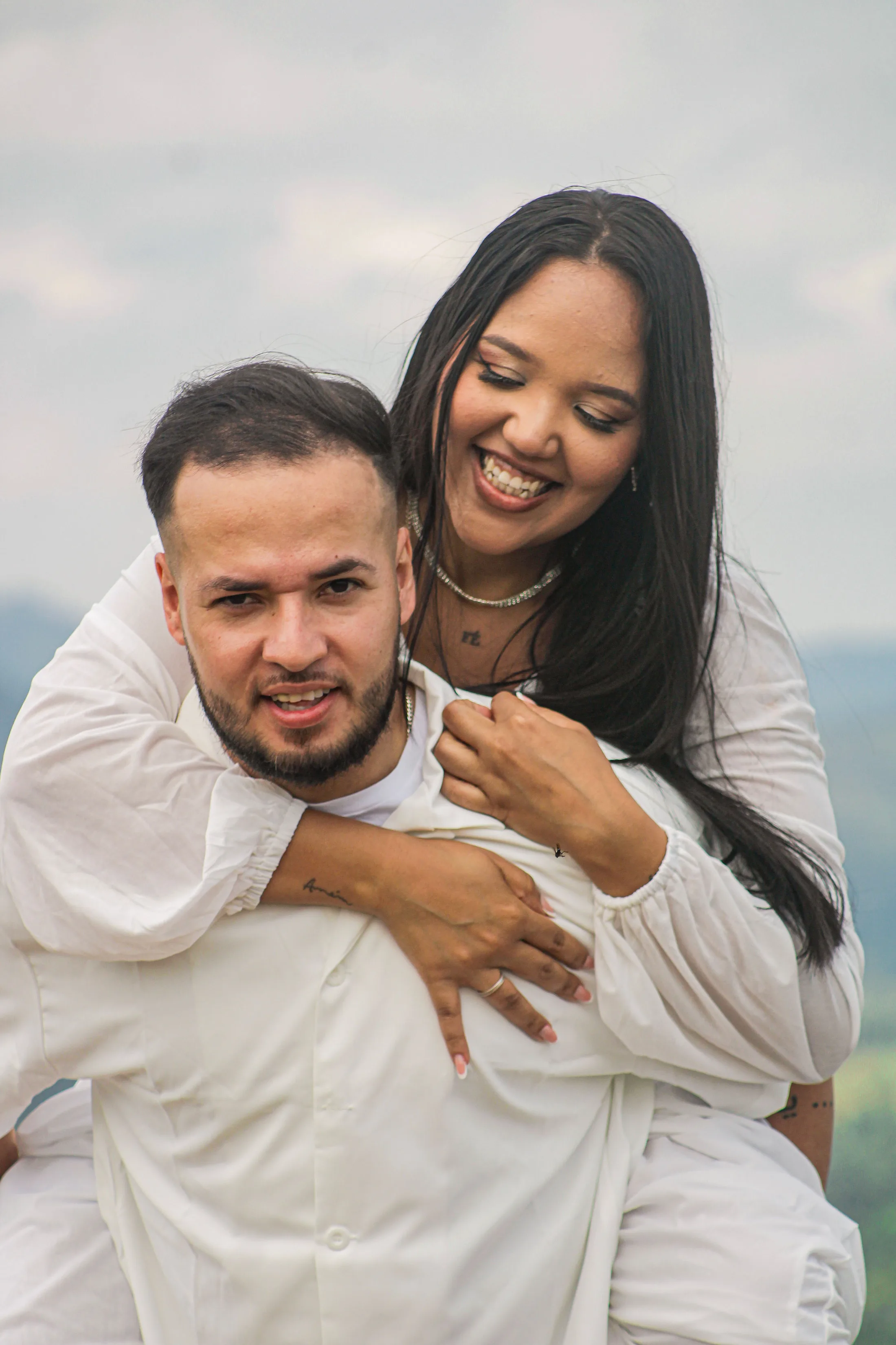 Casal sorrindo; ela o abraça por trás.