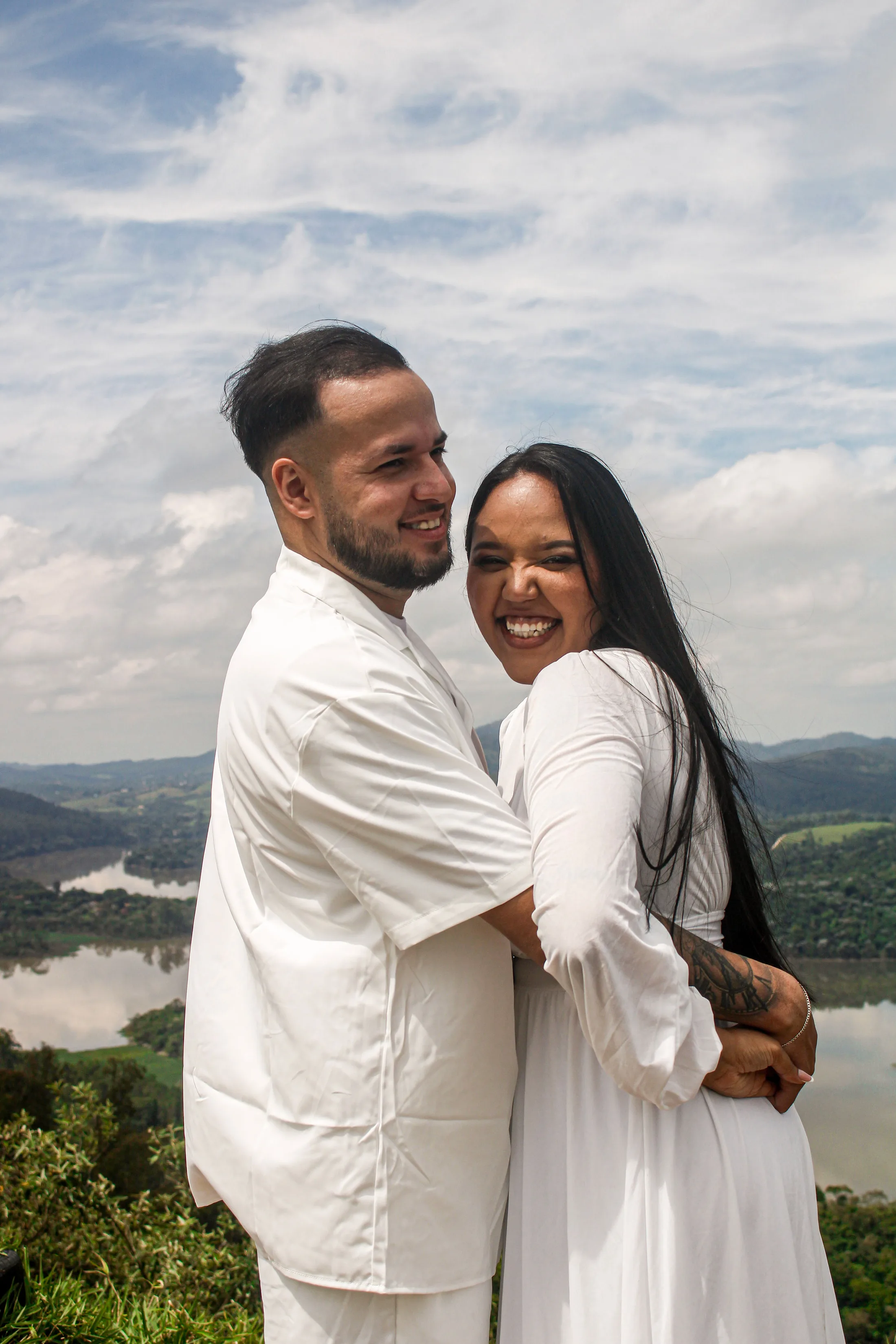 Casal abraçado, sorrindo, com céu nublado ao fundo.