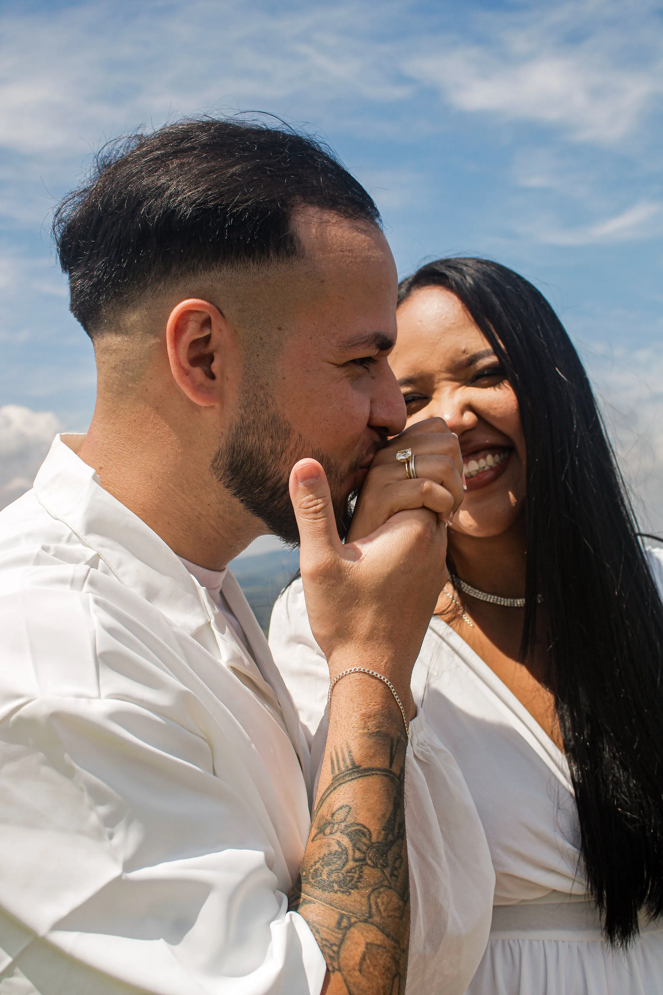 Casal sorrindo; ele beija a mão dela, com céu azul ao fundo.