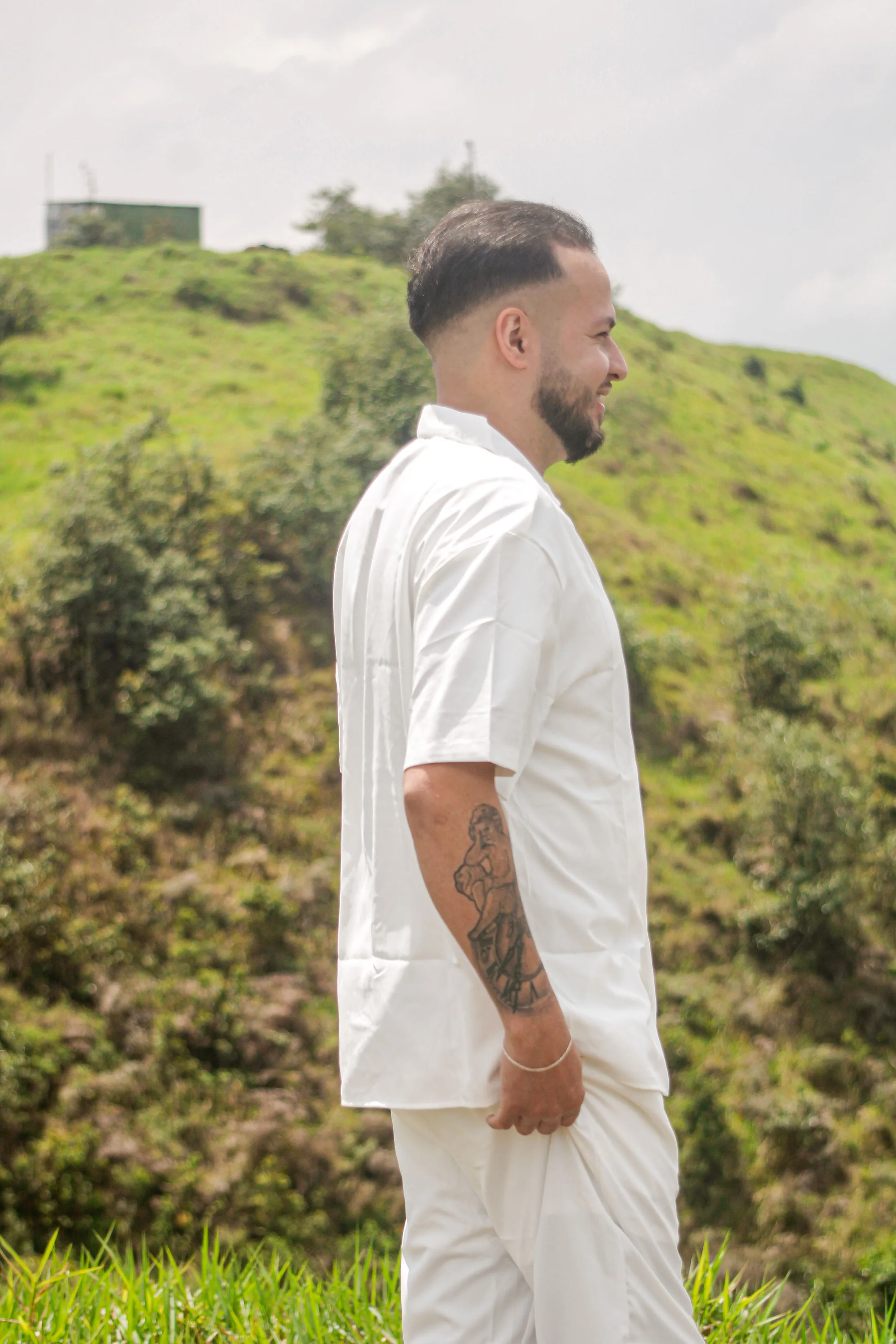 Homem de roupa branca, de perfil, em frente a um morro verde.